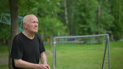 an elderly man tosses a soccer ball away, showing determination and focus, with a blurred view of a goalpost, lush trees, and a building in the background