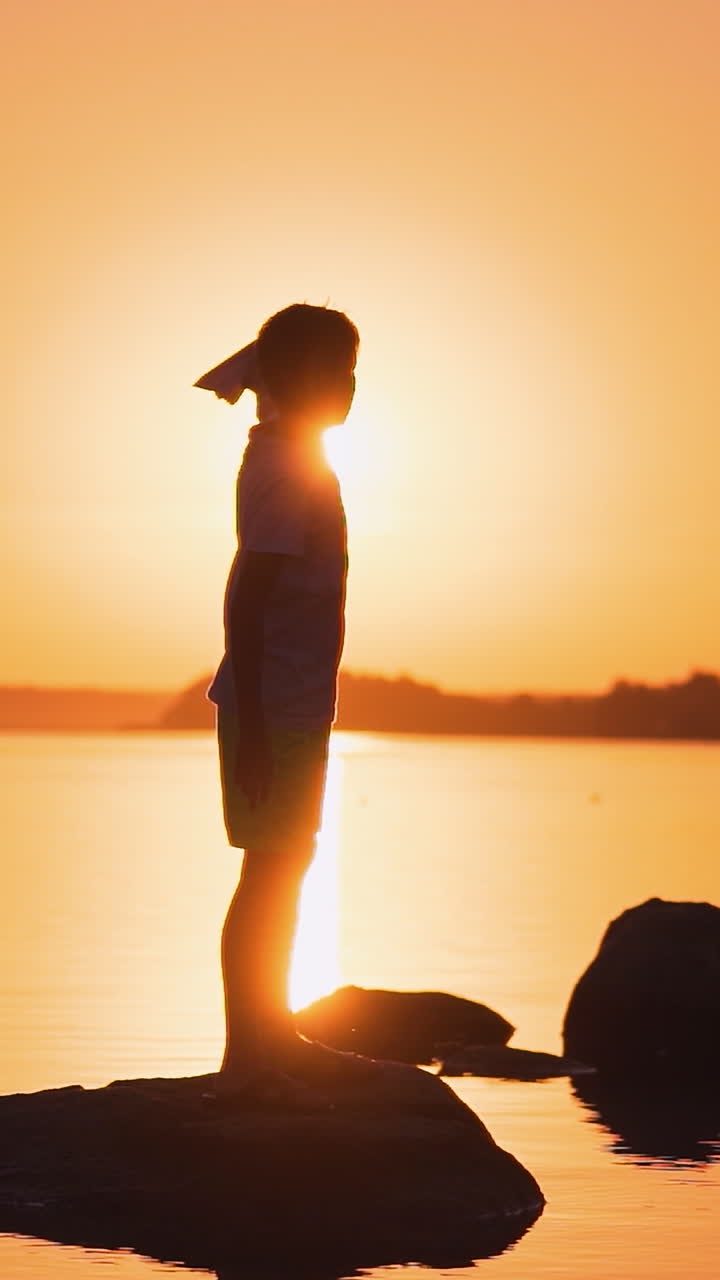 Boy resting near the water. Silhouette of cute young boy standing on stone holding small paper plane in hands Vertical video