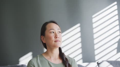 thoughtful asian woman sitting on sofa close to window at home