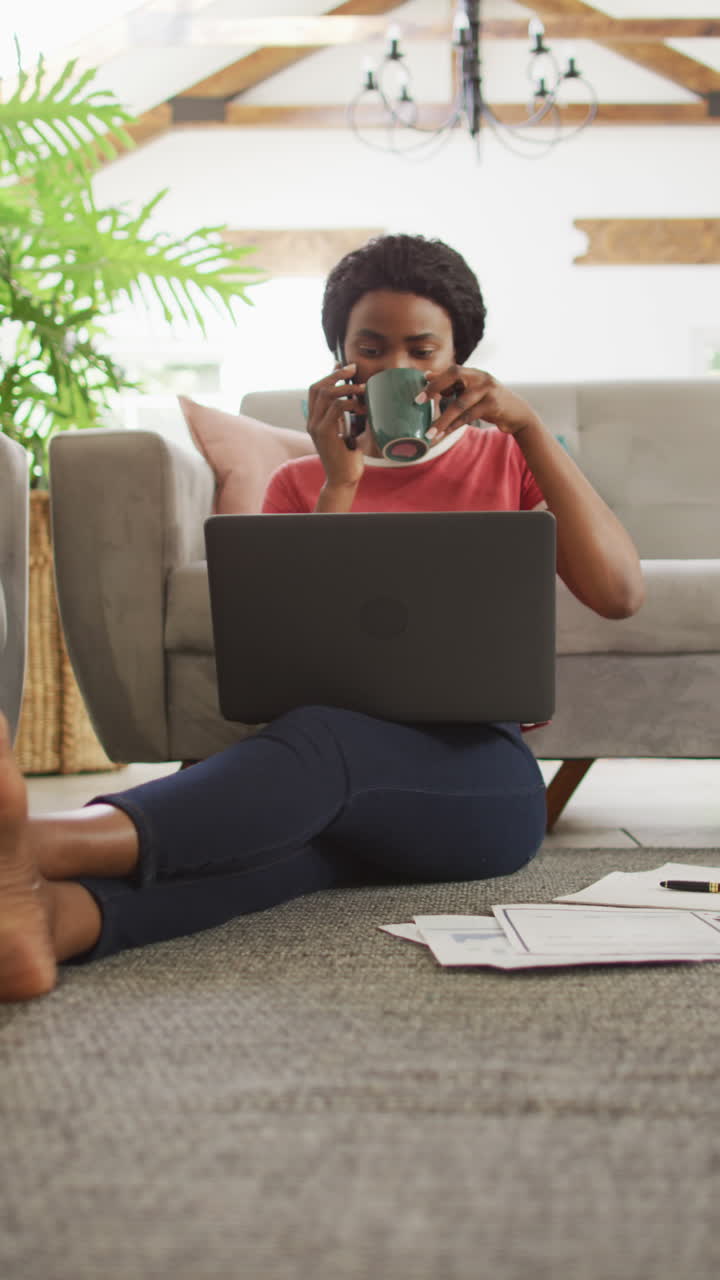 Vertical video of african american woman sitting on floor and using laptop