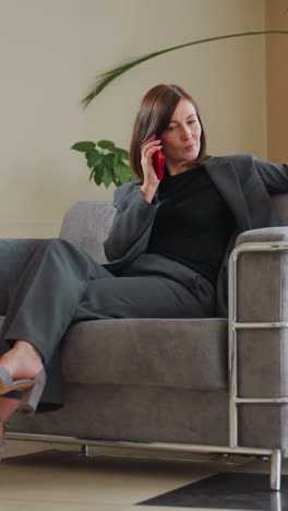 smiling businesswoman in gray suit sits comfortably on modern couch talking on red smartphone in bright office space with large indoor plants, exuding confidence and ease during casual conversation