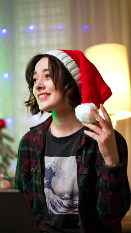 smiling brunette woman in red santa cap looks at her laptop. portrait of a happy pensive lady. christmas tree at backdrop. vertical video.