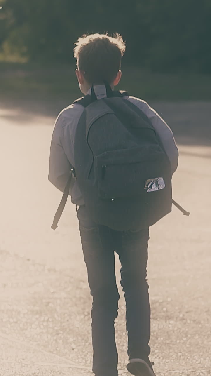 junior schoolboy with bag on back in classic shirt runs along grey asphalt road to green dense trees slow motion backside view