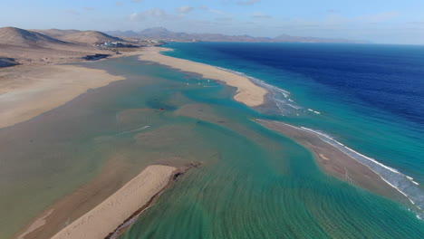 La-Barca-beach,-Fuerteventura:-aerial-view-in-a-circle-over-the-beautiful-beach,-people-practicing-windsurfing-and-kitesurfing