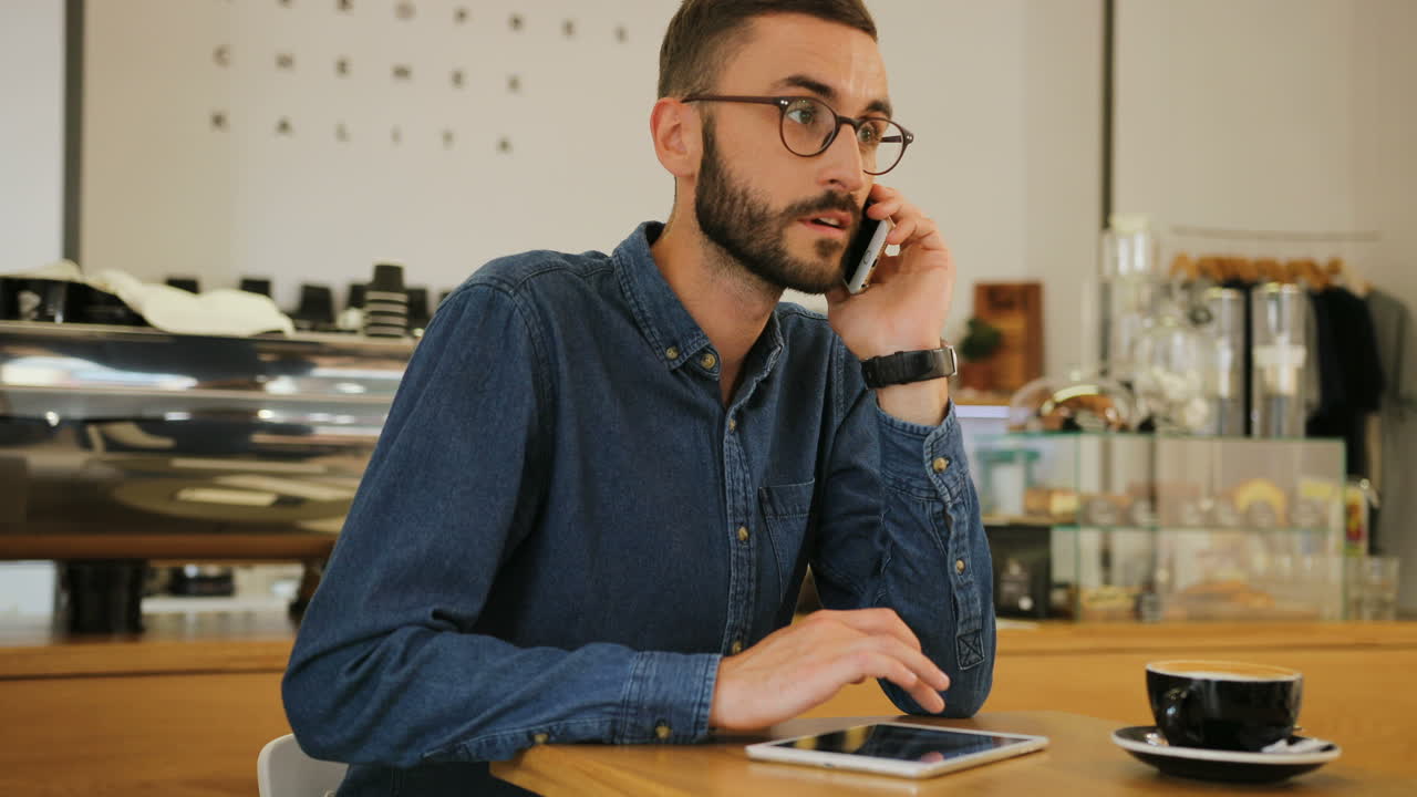 giovane uomo caucasico con gli occhiali che parla sullo smartphone e usa il tablet mentre è seduto al tavolo con una tazza di caffè in una caffetteria