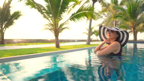 Elegante-Dama-Asiática-Con-Sombrero-De-Verano-Flexible-En-La-Piscina-En-Un-Destino-Tropical