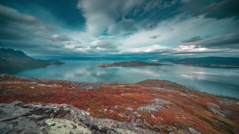 whirling stormy clouds are backlit by the low sun over a calm mirror-like fjord and autumn tundra in the timelapse video