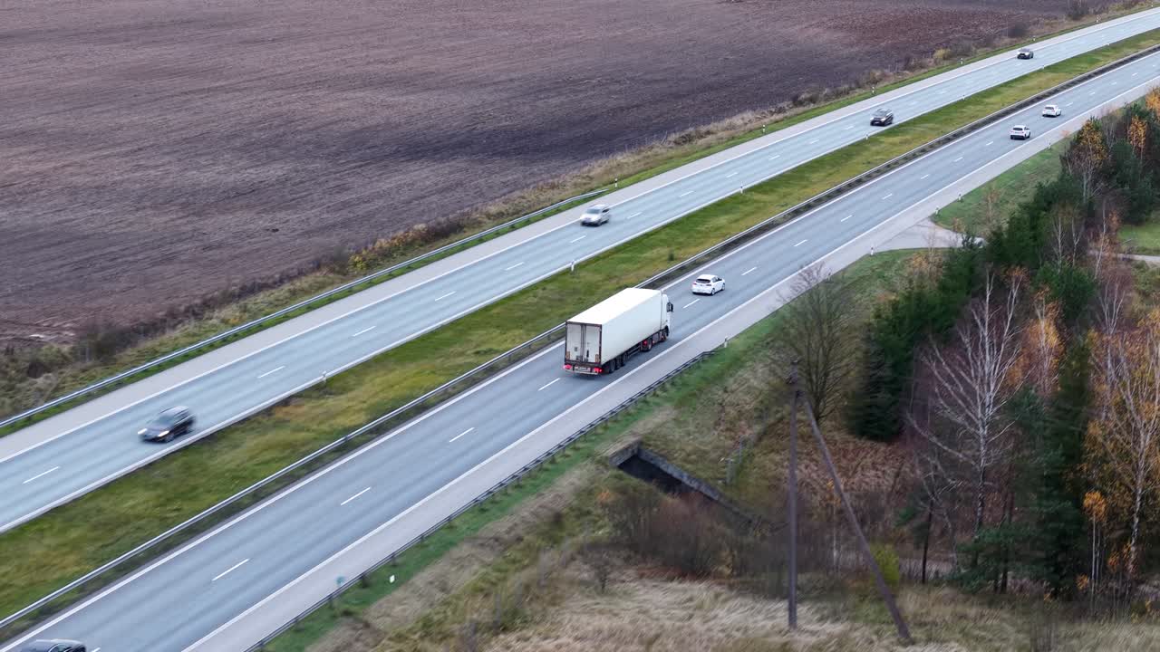 Drone tracks white semi truck driving on Lithuania A2 highway through rural countryside on a cloudy day as cars pass on the left side. Beautiful establishing shot
