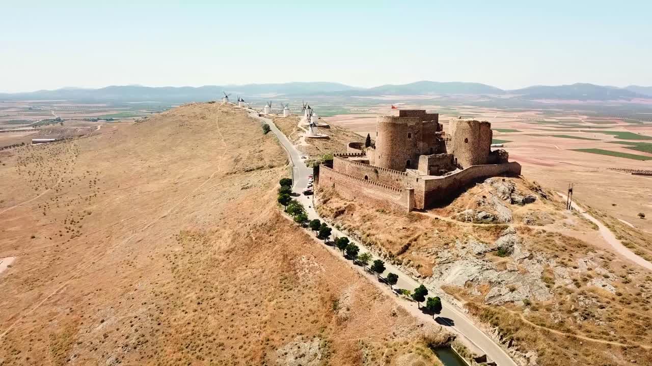 Aerial view of the medieval Consuegra Castle and its iconic windmills standing on a hilltop, overlooking the vast, dry plains of Castilla–La Mancha, Spain, a timeless symbol of Spanish heritage