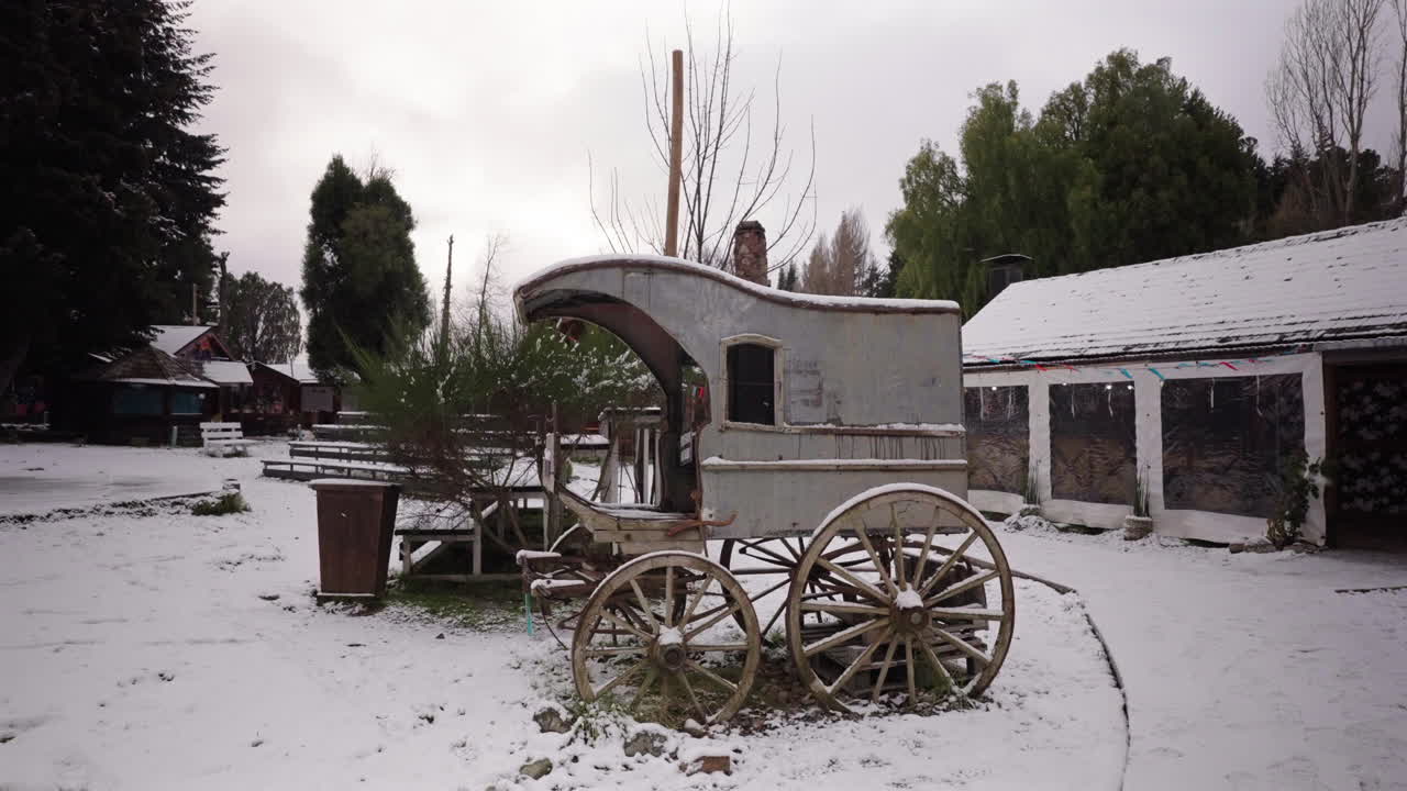 Static camera shots antique wooden horse-drawn carriage covered in snow at Colonia Suiza, Bariloche with traditional Andean setting and wintry landscape evoke rustic heritage charm during snowfall.
