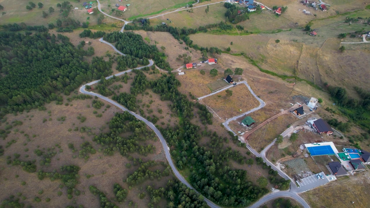 Rural homes and a bright blue pool appear below as the aerial camera rotates over a winding road, fenced plots, dry grassland, and forest edge in Zlatibor, Serbia.