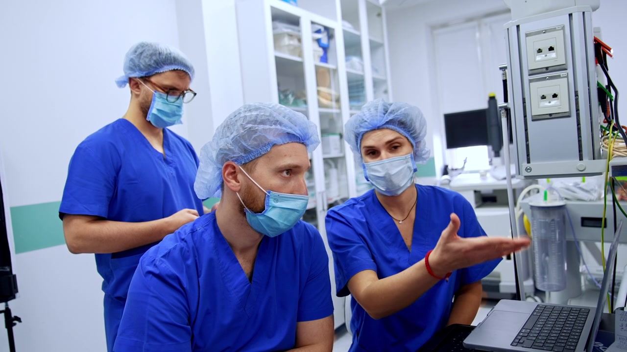 Tres miembros del personal médico con uniformes quirúrgicos azules trabajando en un portátil en un quirófano.
