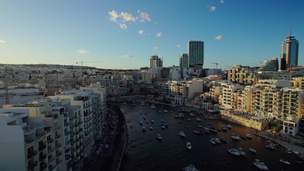 Il drone si muove lungo la baia di St. Julian, mostrando gli edifici illuminati dal sole da un lato e quelli in ombra dall'altro, mentre la città risplende sotto la calda luce del tramonto