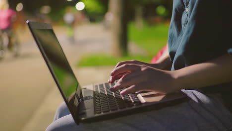 primer plano de una mujer usando una computadora portátil al aire libre en el parque nocturno, dedos escribiendo en el teclado con luces de fondo suaves y personas montando bicicletas