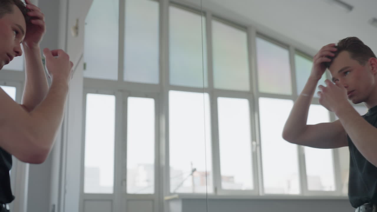 young boy in black vest stands in front of large mirror adjusting hair then turns to pick small black container from chair in bright studio with urban view outside window and soft natural light