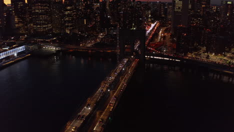 Volar-Sobre-El-Puente-De-Brooklyn-Por-La-Noche.-Vehículos-Que-Se-Mueven-Lentamente-En-Un-Gran-Puente-Sobre-El-Río.-Ventanas-Iluminadas-De-Edificios-De-Gran-Altura-En-El-Paseo-Marítimo.-Manhattan,-Ciudad-De-Nueva-York,-Estados-Unidos