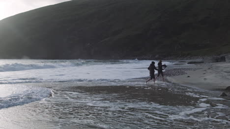 two women running at the beach barefoot on achill island, ireland. waves hitting the beach. handheld slow-motion shot with huge hill in the background.