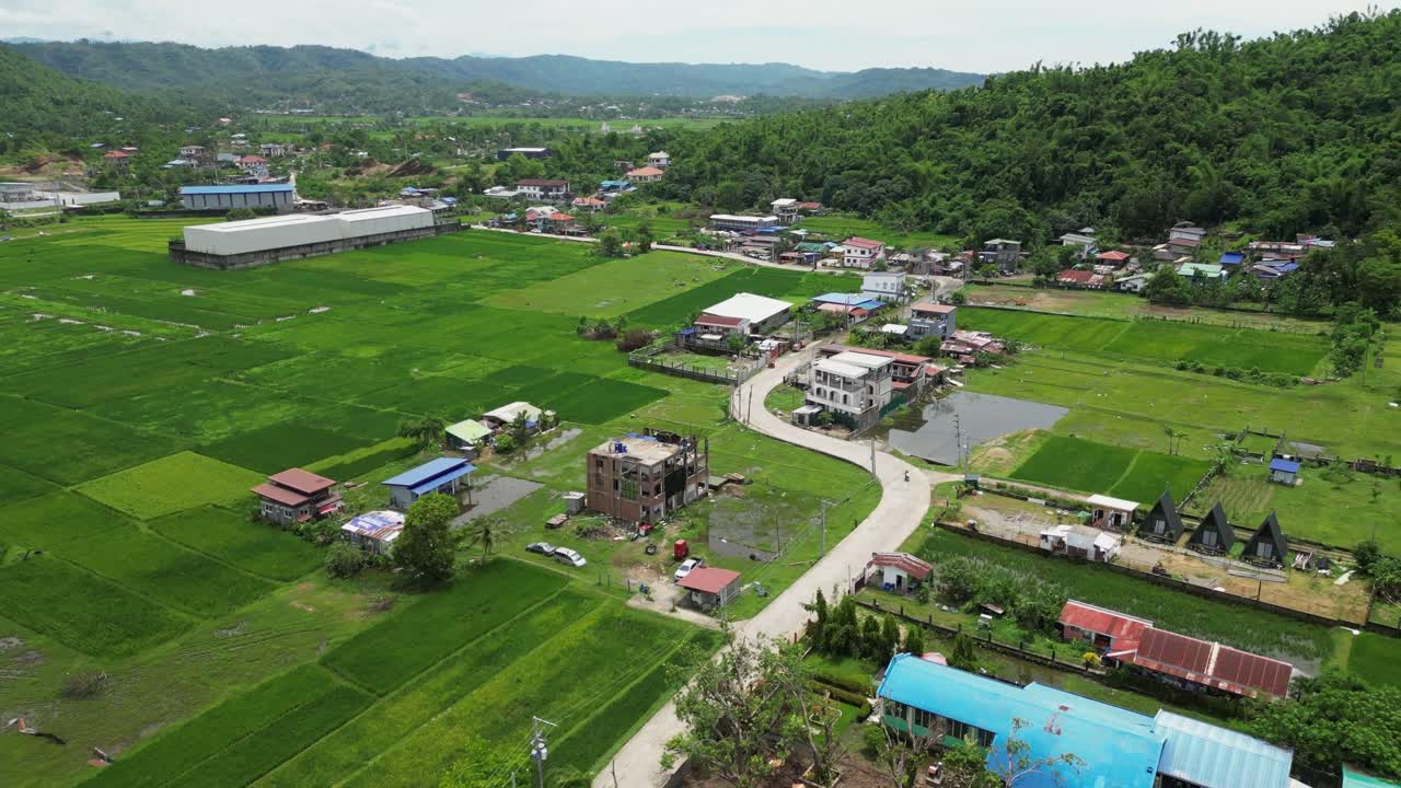 Wide aerial footage reveals a sprawling countryside of rice fields, clustered rooftops, and winding roads, framed by forested mountains and lush agricultural terrain
