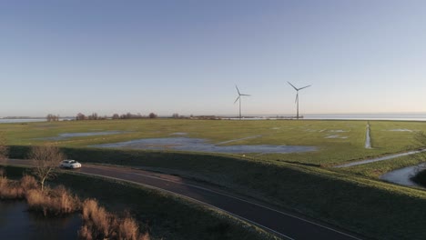 Luftaufnahme-Eines-Weißen-Autos,-Das-Bei-Sonnenuntergang-Auf-Einer-Kleinen-Landstraße-Mit-Zwei-Windmühlen-Im-Hintergrund-Fährt