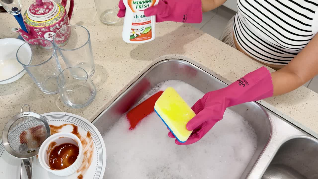 Person wearing pink gloves dispenses liquid soap onto a yellow sponge at a kitchen sink with dirty dishes, under bright indoor lighting, top-down view