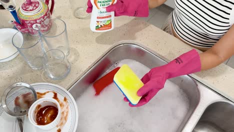 person wearing pink gloves dispenses liquid soap onto a yellow sponge at a kitchen sink with dirty dishes, under bright indoor lighting, top-down view