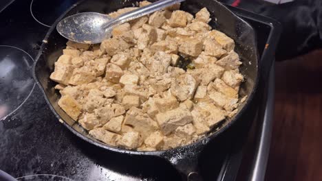 Kochen-Von-Veganen-Tofu-Würfeln-Auf-Pflanzlicher-Basis-Aus-Soja-Auf-Einem-Schwarzen-Herd-Und-Einer-Gusseisernen-Pfanne,-Während-Die-Kamera-Zurückfährt-Und-Jemand-Mit-Der-Gabel-Proben-Aus-Dem-Topf-Nimmt-–-In-4K,-Halbe-Geschwindigkeit,-30-Bilder-Pro-Sekunde