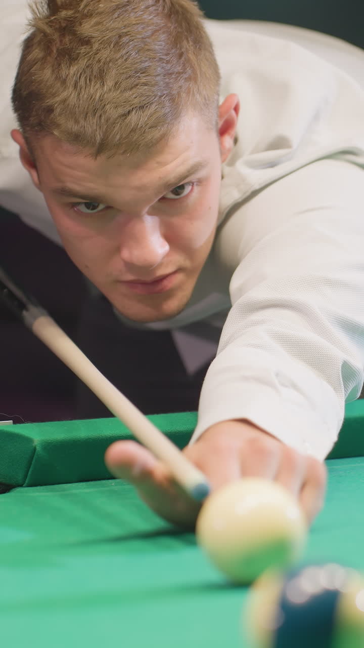 vista frontal de primer plano del hombre del juego en camisa blanca con cara seria, inclinado sobre la mesa de billar verde en una habitación débilmente iluminada. agarrando el palo de señal con enfoque, listo para golpear la bola blanca