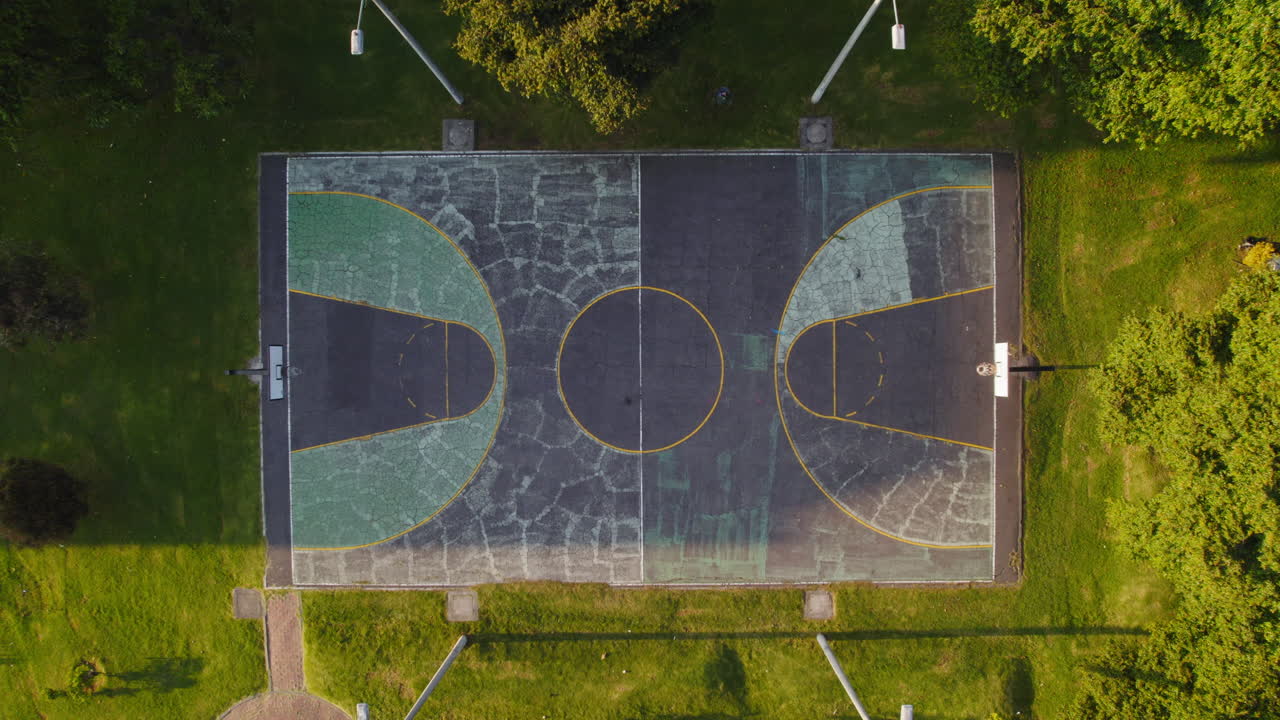 Vista aerea con drone di un campo da basket vuoto in cemento crepato in un parco all'aperto, circondato da erba verde lussureggiante e alberi, durante un'alba o un tramonto soleggiato.