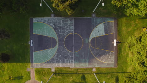 aerial drone view of cracked concrete empty basketball court in outdoor park surrounded by lush green grass and trees during sunny sunrise sunset