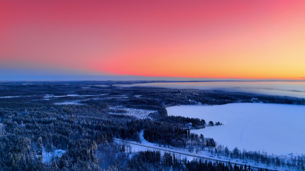 Vidt luftfoto av frossen innsjø og skog ved solnedgang. Panoramisk droneopptak av en snødekt skog og frossen innsjø i Lappland under en levende rosa og oransje solnedgangshimmel.