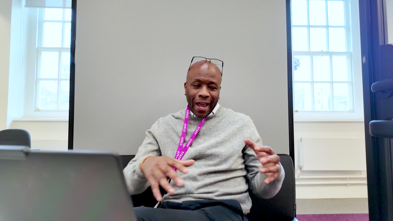Black Businessman wearing gray sweater and lanyard gesturing with hands while speaking inside office pod