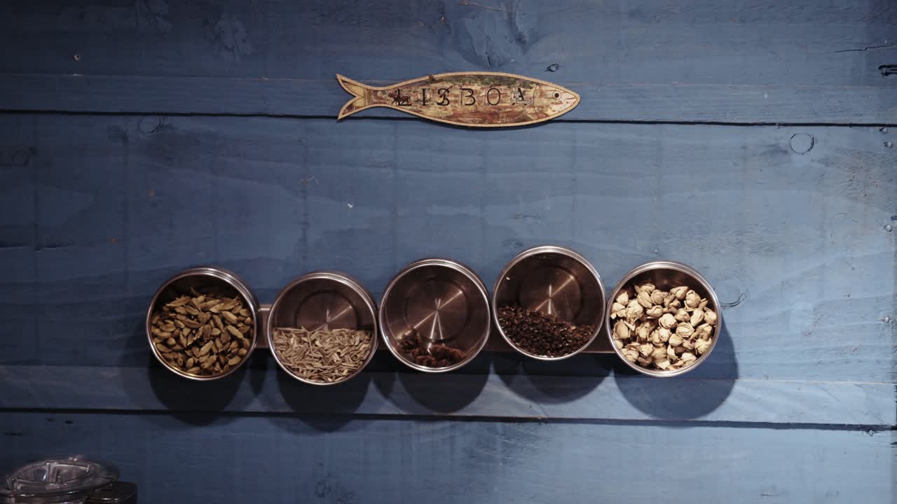 Fünf Schalen gefüllt mit verschiedenen Gewürzen, begleitet von einem Fischsymbol, auf blauem Holzuntergrund, die natürliche Aromen und Kochkunst symbolisieren.