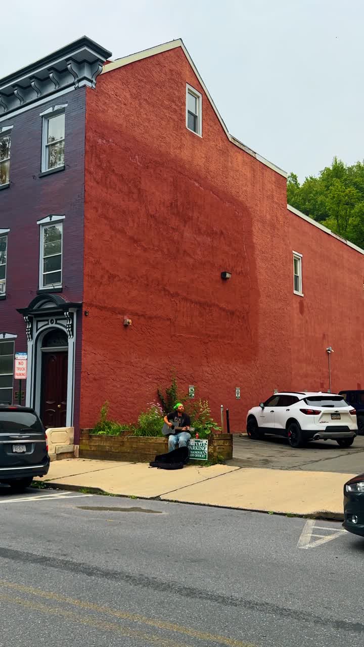Jim Thorpe, USA, 2 luglio 2025: Artista di strada che suona la chitarra acustica fuori da un edificio di mattoni rossi. Uomo seduto su un muro di pietra che suona la chitarra in un parcheggio urbano con uno sfondo di parete rossa.