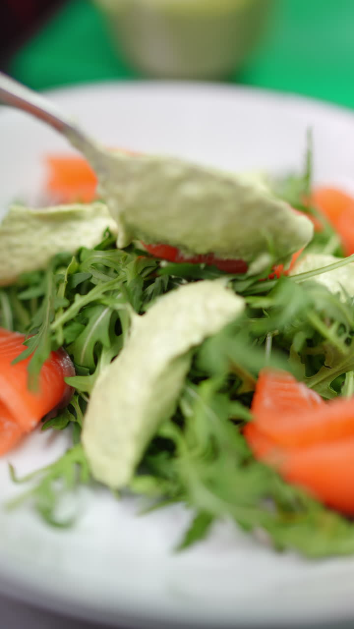 Vertical footage Green sauce is being added with a spoon on a fresh salad with arugula, salmon and tomatoes on a white plate and green background