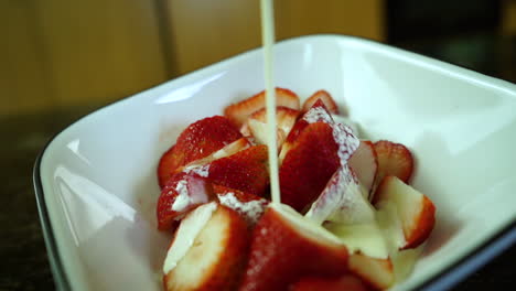 crema batida pesada goteando lentamente sobre fresas maduras en un tazón blanco en un mostrador de cocina, cámara lenta de primer plano
