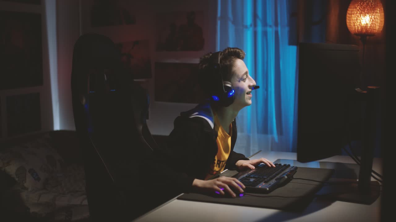 Young Caucasian Guy With Headset, Talking While Playing Online Game On The Computer Inside The Room - medium shot, side view