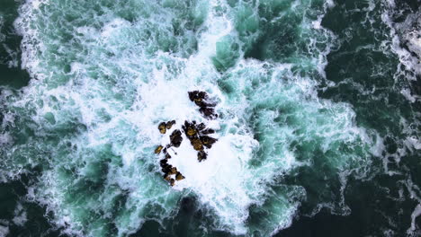 Aerial---Drone-rises-from-rock-in-ocean,-incessant-pounding-of-waves