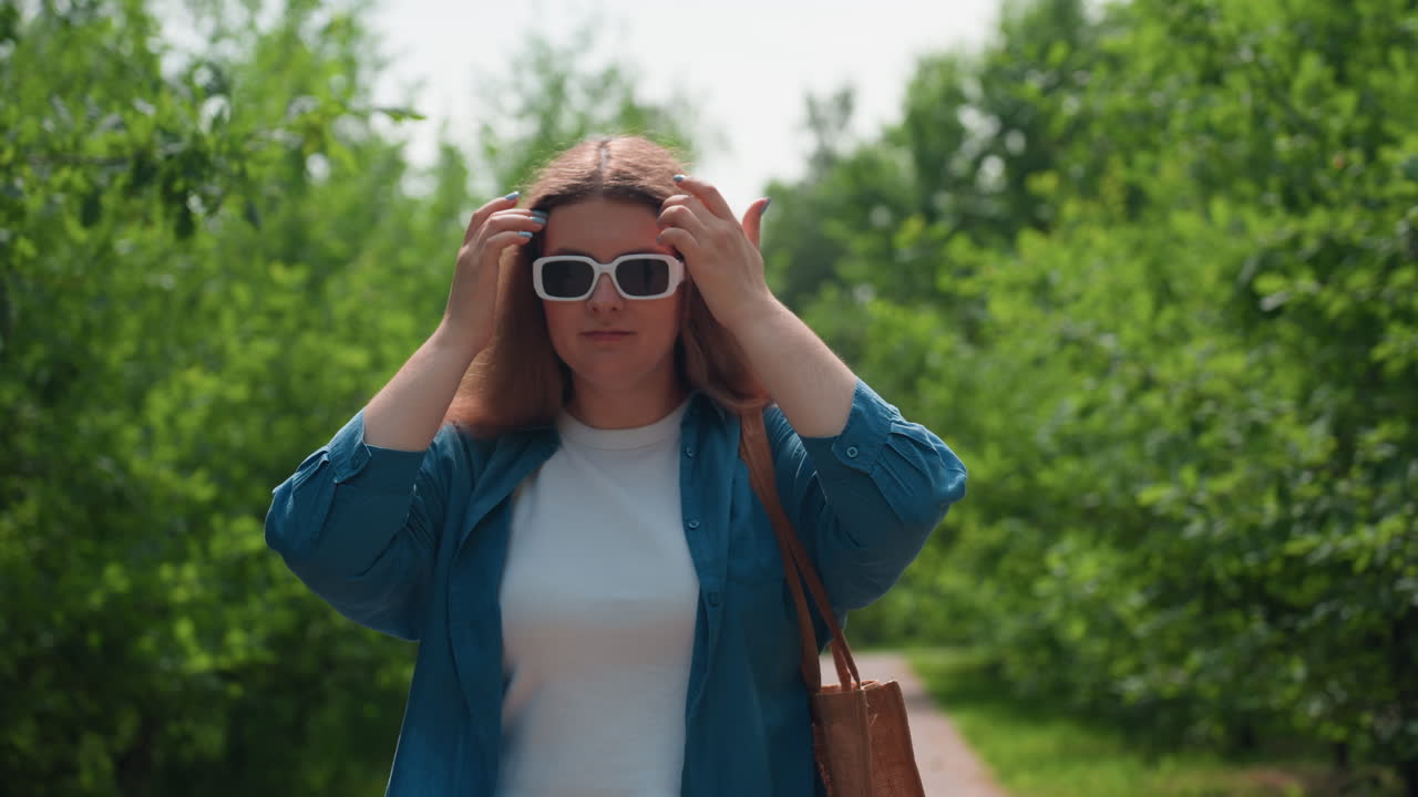 Молодая женщина в солнечных очках, стоящая на улице в окружении зеленых деревьев, спокойное уверенное выражение лица в летнем солнечном свете, синяя рубашка и белый наряд, расслабленное настроение