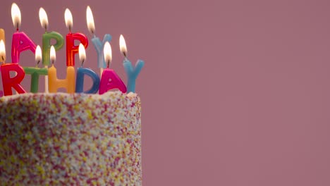 hand lighting candles spelling happy birthday on cake covered with decorations against pink studio background