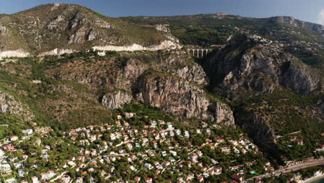 Aerial-flyover-small-french-village-on-mountain-during-sunny-day-with-blue-sky-and-bridge-in-background-between-rocks