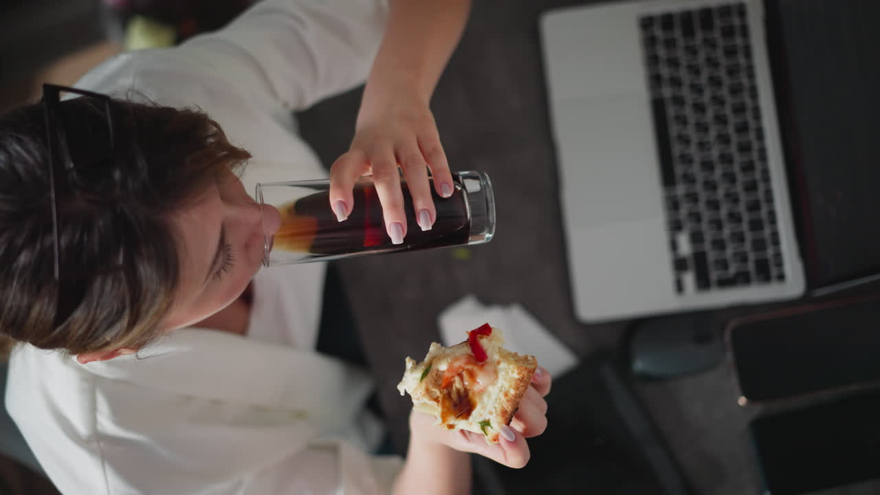 Aerial view of woman wearing white shirt drinking juice and eating pizza slice while sitting at cluttered office desk with laptop, smartphone, computer mouse, and papers during busy work break