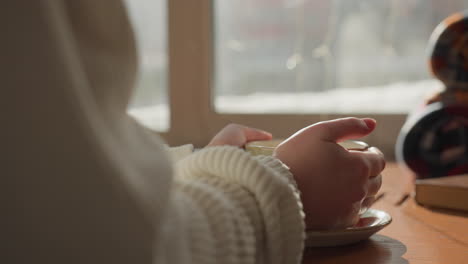 close up of individual in white woven sleeve gently holding warm coffee cup on wooden table as soft sunlight pours through window, casting reflections and shadows in calm morning setting