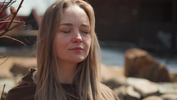 Close view of young woman with closed eyes and soft smile, holding relaxed mindful stillness on rocky shoreline while warm golden sunlight highlights freckled skin, long hair, and peaceful outdoor