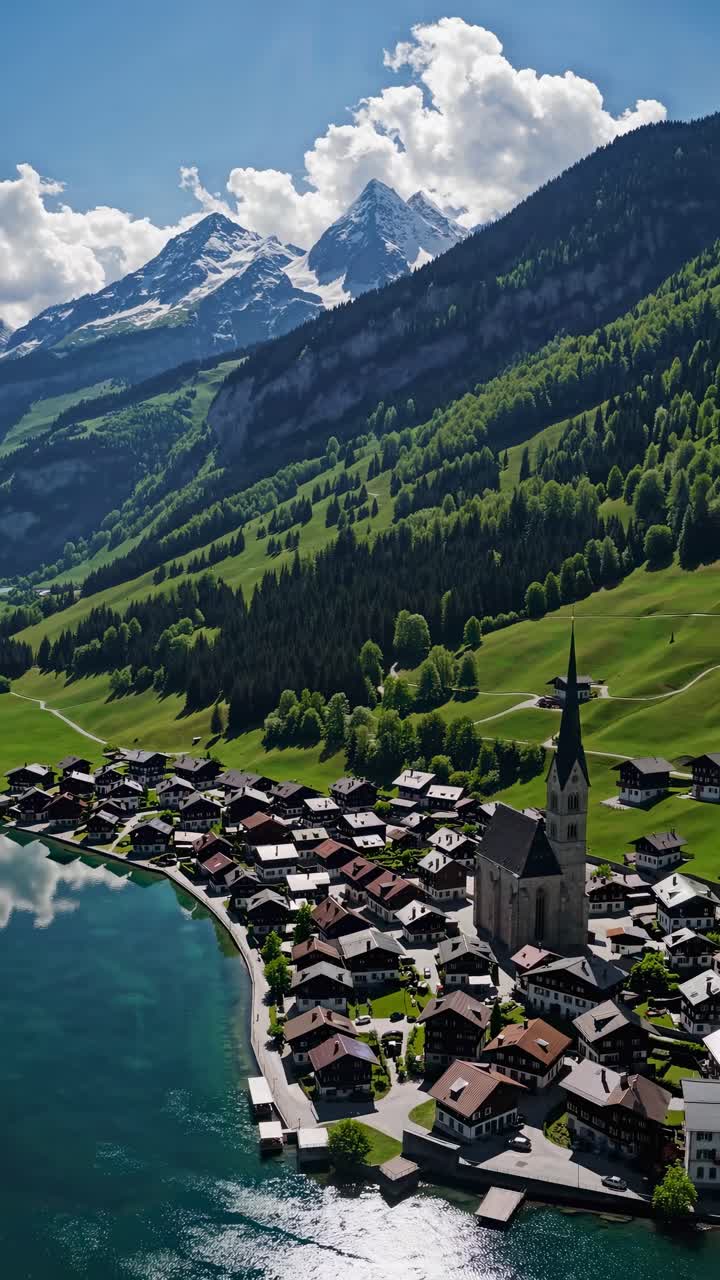 Aerial video captures a serene alpine village by a lake, framed by lush hills and majestic