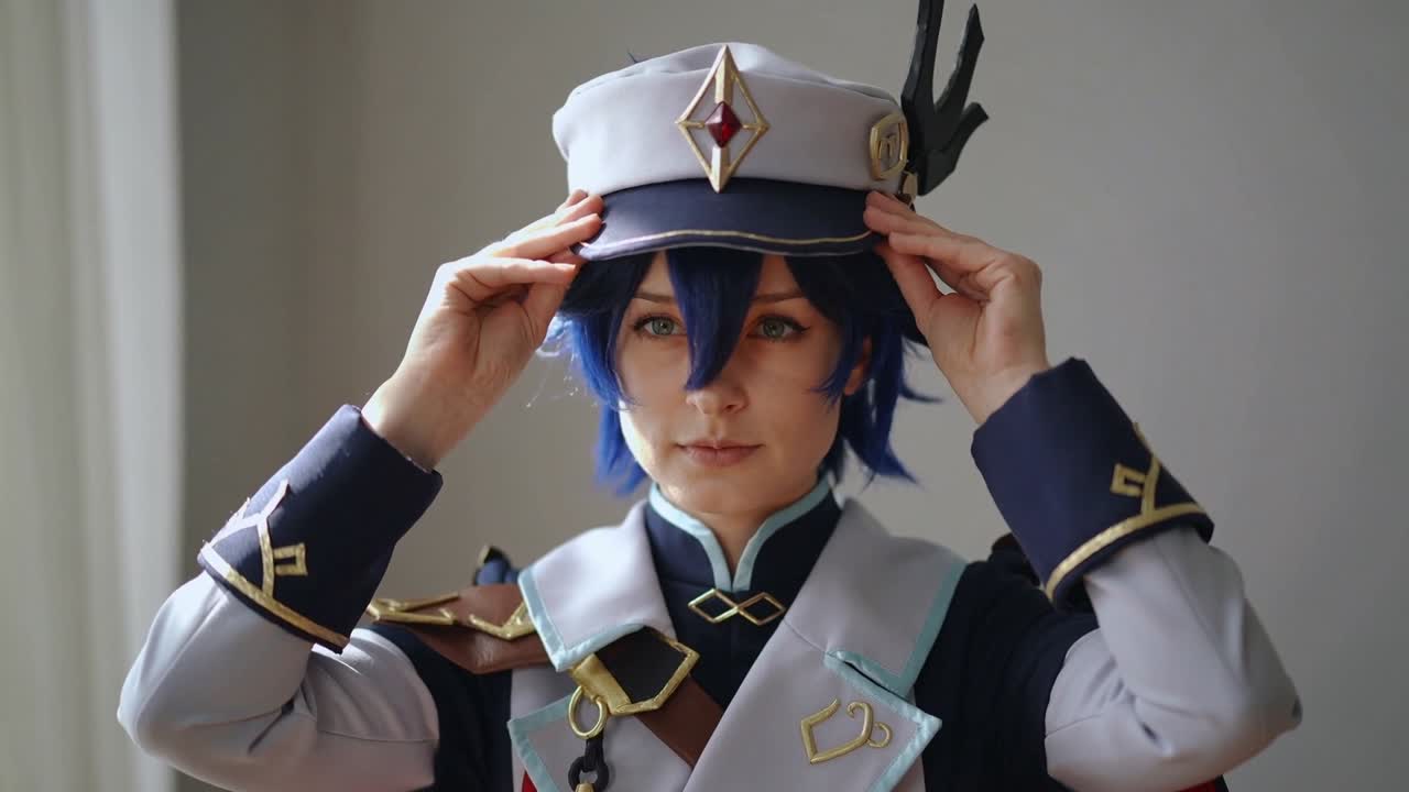 スタジオで、制服姿のコスプレイヤーがつばを押さえながらつば付きの帽子を持ち上げ、青いウィッグを見せてポーズを準備している。