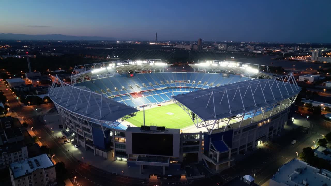 Panoramująca kamera drona przesuwa się w zanikającym świetle zachodu słońca, ukazując oświetlone boisko stadionu i miasto.