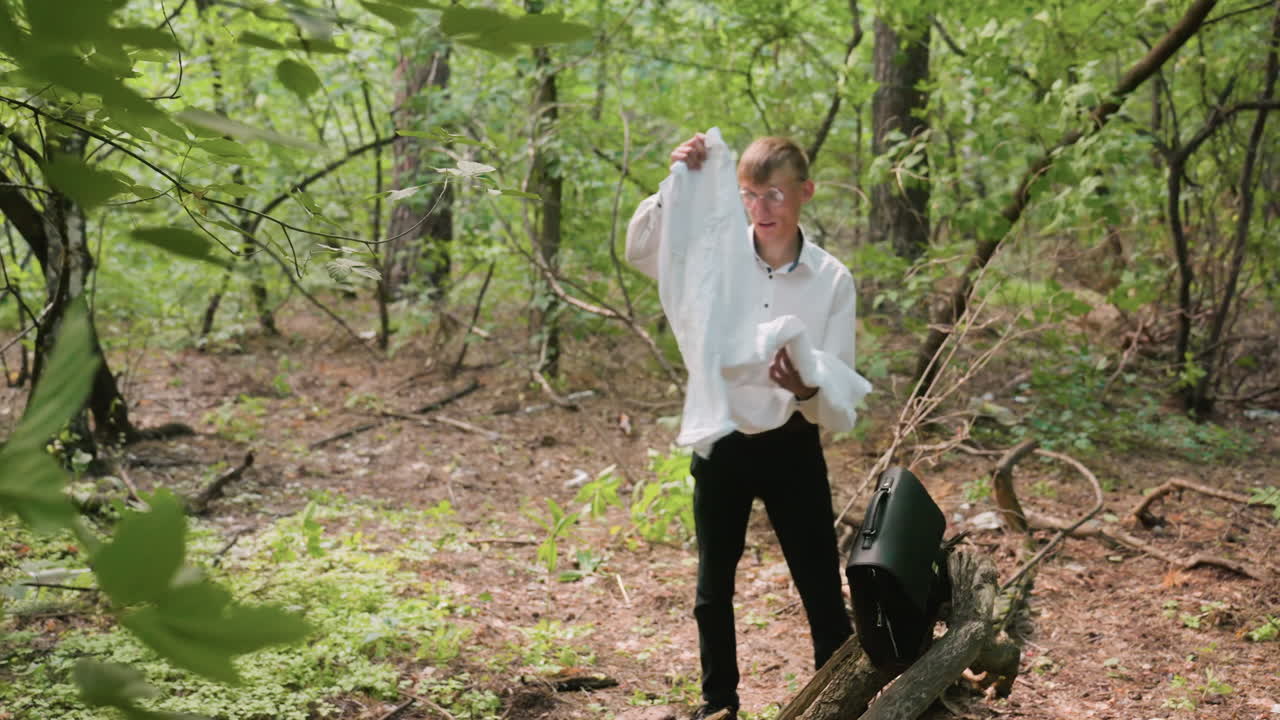 Rear view of man in white shirt bending over in forest while removing white coat from black leather bag placed on ground, surrounded by green foliage, trees, and natural woodland setting
