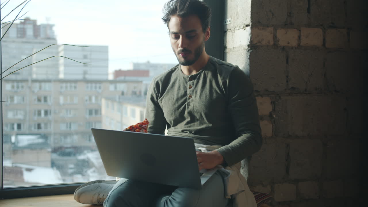 Man Eating Pizza and Working on Laptop by the Window