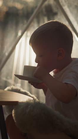 child takes sip of tea at low table at glamping. little boy drinks hot beverage sitting on floor in luxury camp. kid enjoys calmness on vacation