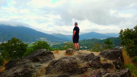 hombre caminando desde la cima de la montaña después de disfrutar de la vista de un valle lleno de casas y rodeado de montañas cubiertas de nubes, cámara lenta